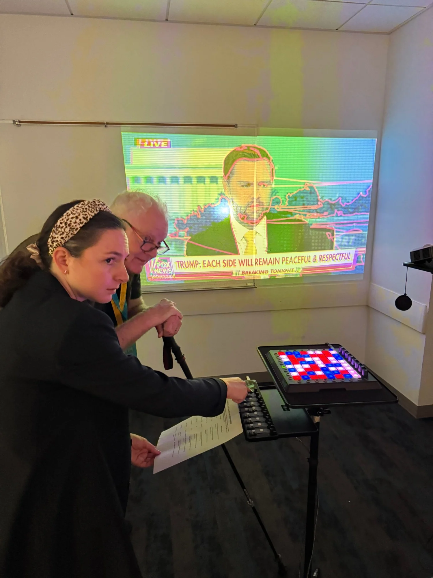 Two visitors at the fader bank, a news broadcast traced in red and recoloured on the wall behind them